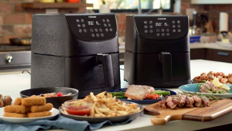 Two Cosori air fryers with plates of food in front of them sit on a kitchen counter.