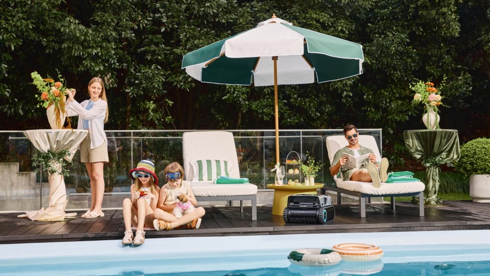 A young family relaxes by the pool under an umbrella while the Aiper Scuba X1 Pro Max cleans the pool nearby