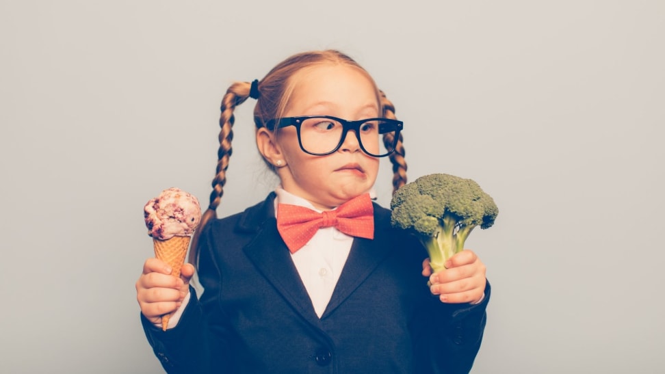 Little girl holds ice cream in one hand, broccoli in the other hand, making a silly face at the broccoli.