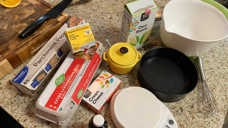 Various ingredients are laid out on a countertop: corn starch, whipping cream, large brown eggs, cream cheese, and so on.