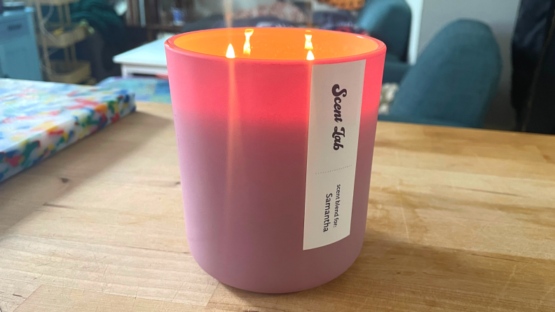 A lit, personalized pink Scent Lab candle on a wooden table in a bedroom.