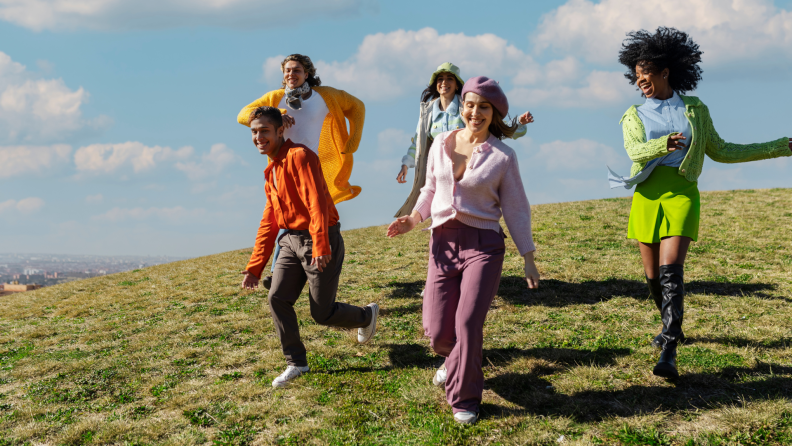 A group of stylish friends joyfully running down a grassy hill on a sunny day.