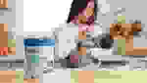 Mother holds baby and feeds with a bottle in the background while Nara Organics formula container sits on counter in foreground