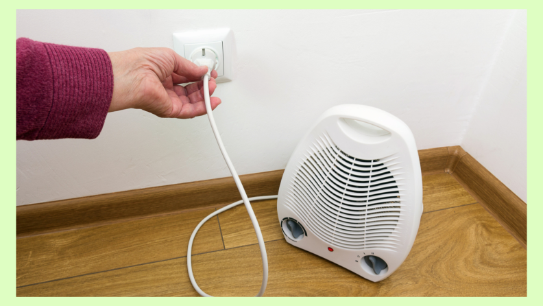 Small white space heater placed on ground with hand plugging the heater into a wall socket