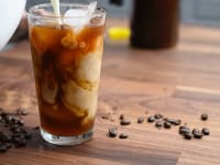 Milk pouring into a glass of cold brew coffee  surrounded by coffee beans sitting on a wood counter.