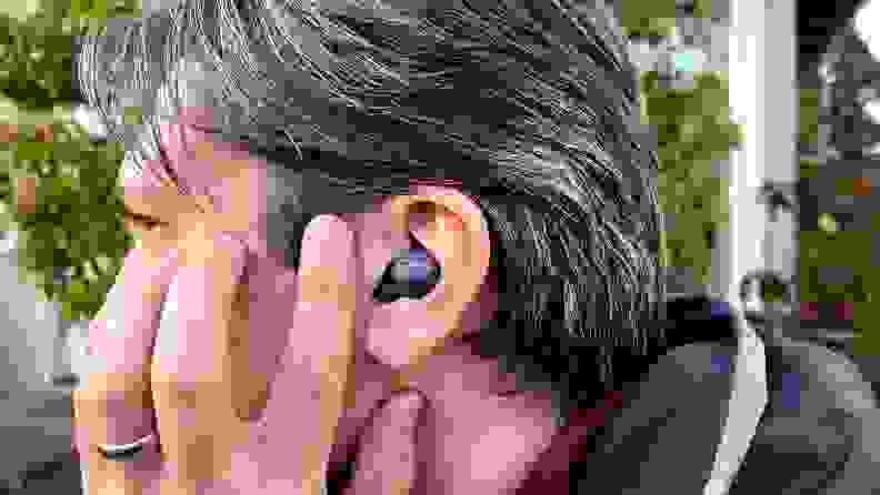 A man wearing a gray coat with salt and pepper hair taps his cheek in front of a grey, double looped earbud.