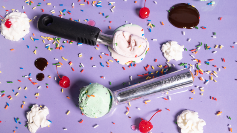The OXO and Zeroll scoops filled with ice cream, laying on a purple background surounded by  sprinkles, cherries, and ice cream.