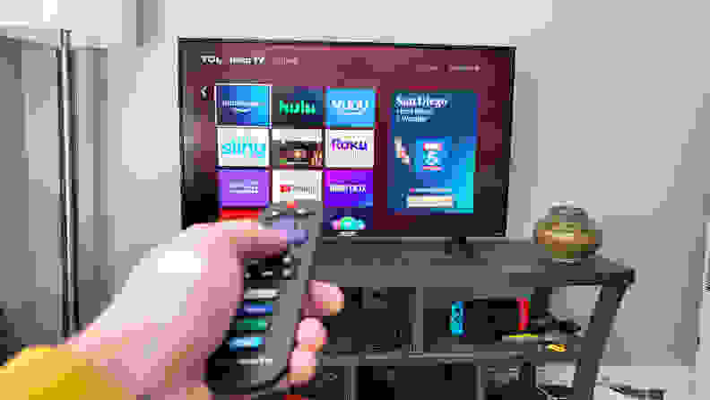 A person pointing a Roku remote at a TV mounted in a living room showing the Roku home screen.