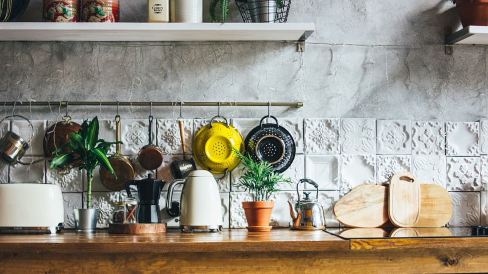 A sunlit kitchen counter filled with plants and small appliances including a black moka pot, wooden cutting boards, a white two-slice toaster, a white electric kettle, and two colanders hanging above them on the textured white wall.