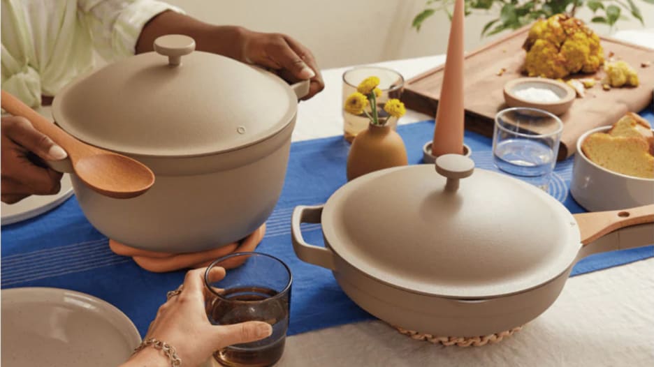 A pair of hands holding a beige Our Place pot and placing it on a table with a similar colored skillet.