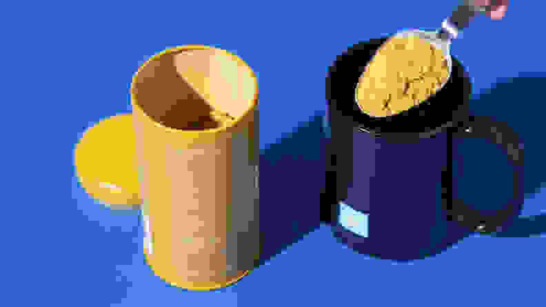 Mud/Wtr product containers, in the color blue and yellow, with the lid off and a scoop of powder being removed from the container.