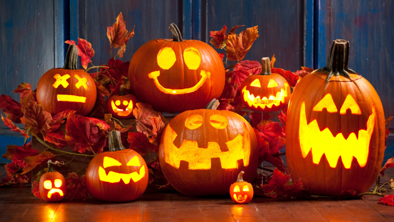 Illuminated Jack-O-Lantern pumpkins of different sizes gathered together surrounded by fall leaves on top of wooden floor.