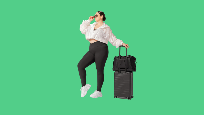 A model wearing black tights, a white jacket, toting around a black suitcase.
