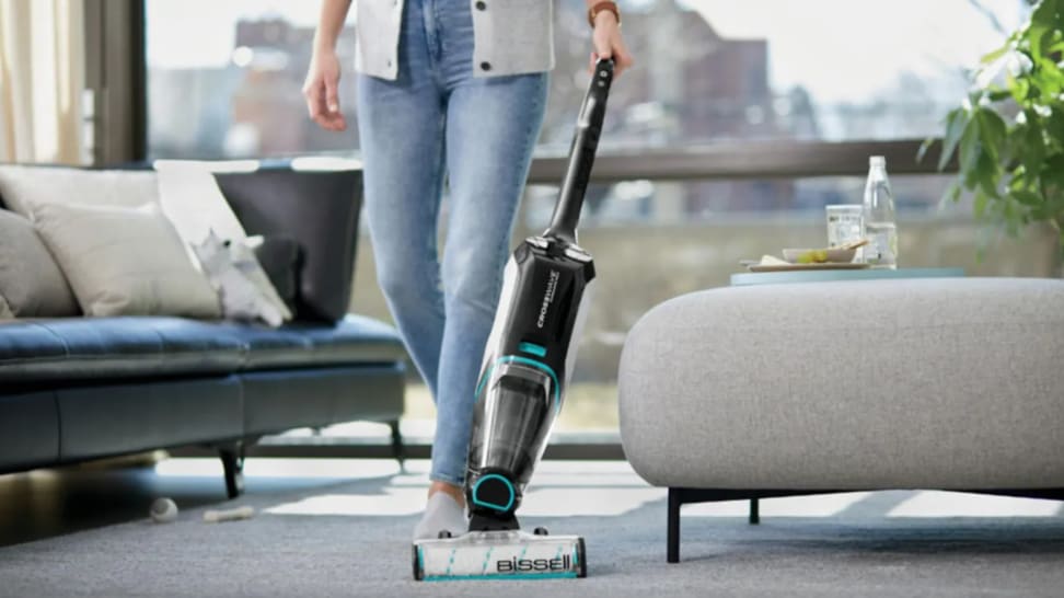 Person using vacuum to clean carpeted floors in living room at home next to couch.