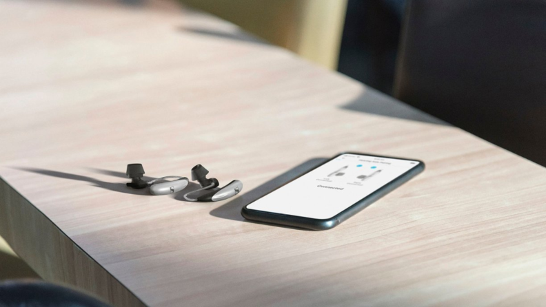 The Sennheiser All-Day Clear hearing aids sitting on a table next to a smartphone