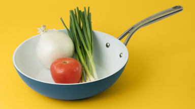 In a green Frök frying pan with ceramic coating, there's a tomato, an onion, and a bunch of spring onions.