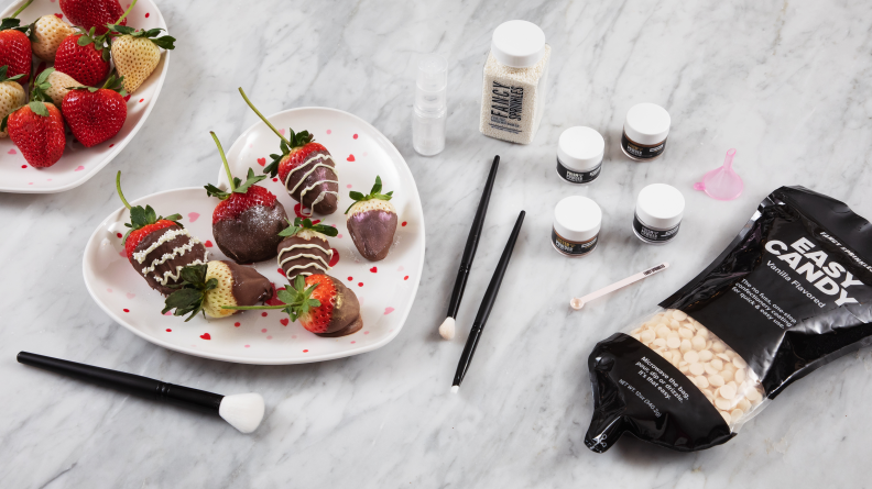 Chocolate covered strawberries on a heart-shaped plate, brushes, pots of edible glitter, and a bag of white chocolate chips sit on a marble counter.