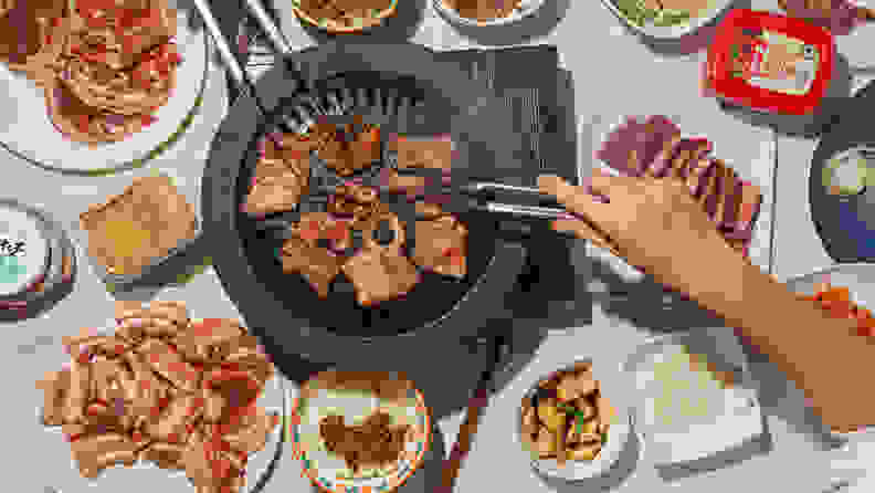 A typical Korean barbecue spread with a iron barbecue stove in the middle, grilling sliced pork belly. Around the stove, there are plates of thinly sliced meat, pickled vegetables, and sauces.