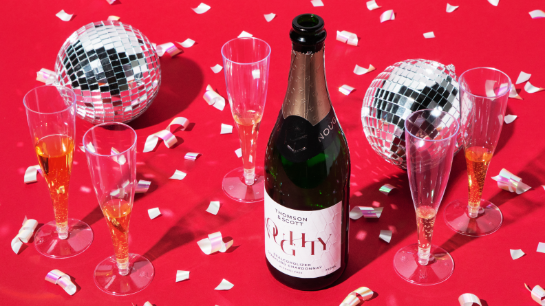 A bottle of Noughty Alcohol-Free Sparkling Chardonnay on a red background with confetti and disco balls.