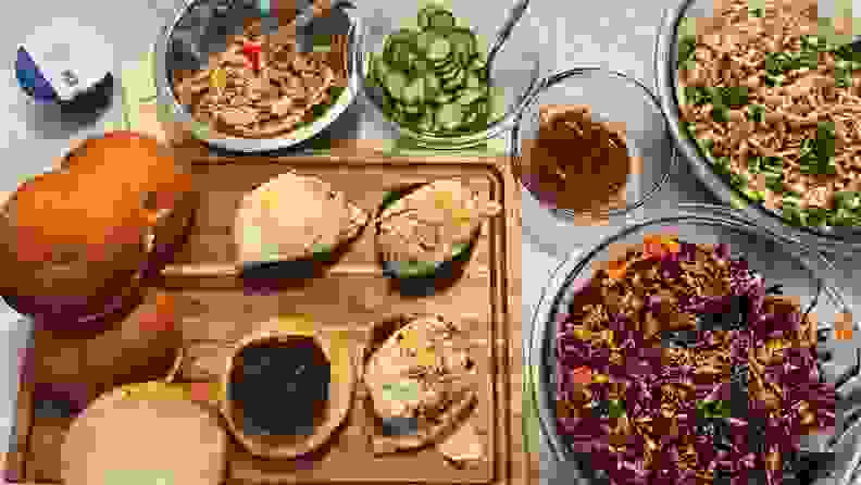 Burgers and buns on a wood cutting board surrounded by condiments, cabbage slaw, and peanut noodles in glass bowls.