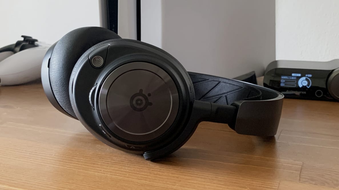 The SteelSeries Arctis Nova Pro Wireless gaming headset sits in front of its base station and a PlayStation 5 console on a wooden table.