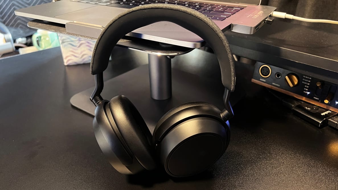 A pair of black headphones with a fabric-topped headband sit on a grey laptop on a black desk with gear behind it.