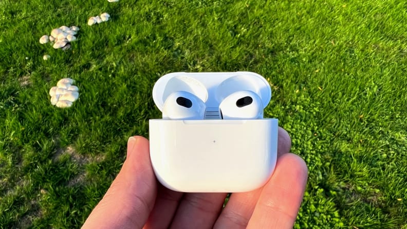 The all-white AirPod sits in their case, held before a lush green field with mushrooms growing to the left.