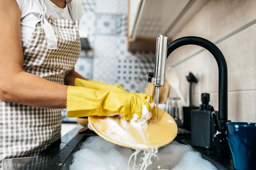 A person wearing a gingham apron and yellow rubber gloves is standing at the sink washing dishes.