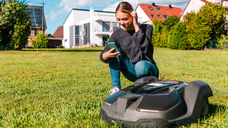 A person holding a smartphone kneels down next to a robot lawn mower on a large grassy lawn outdoors on a bright, sunny day.