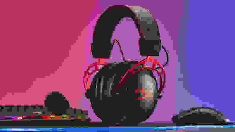 A black gaming headset with red trim sits on a desk next to computing peripherals with a rainbow backdrop.