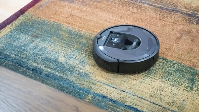 An iRobot Roomba on a green and blue patterned rug, vacuuming up crumbs.