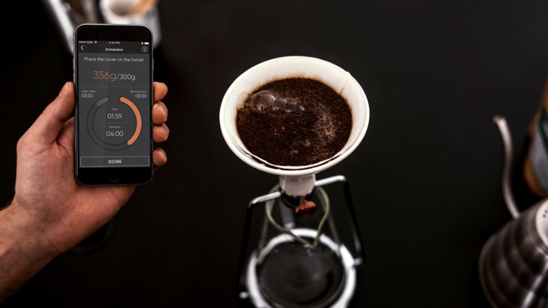 A person is holding a smartphone with Gina coffee maker's app displaying brewing data. Besides the person, there's a Gina coffee maker making pour-over coffee.