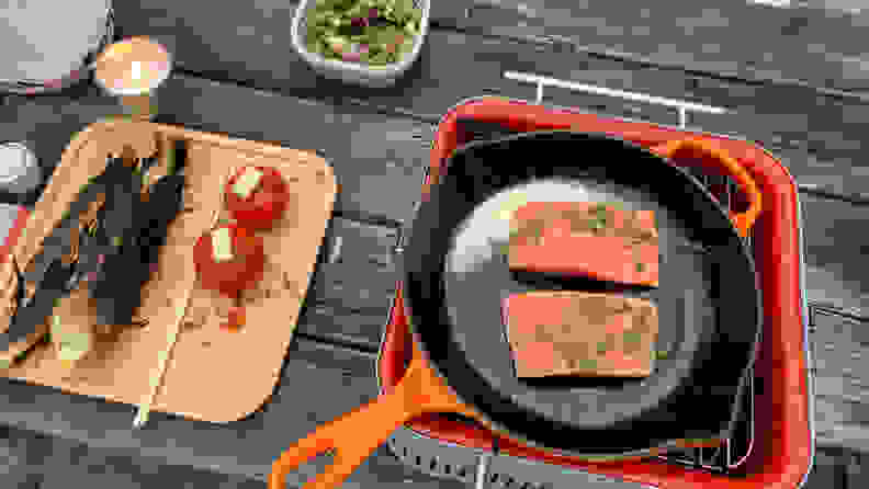 An orange cast iron skillet is placed on top of the Everdure Cube charcoal grill. In the skillet, there are two pieces of salmon. Next to the grill, there's a bamboo tray of cooked corn and tomatoes.