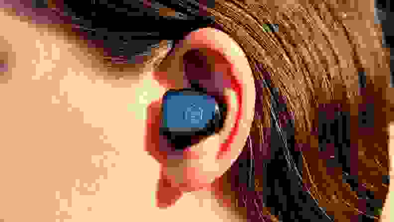 Master & Dynamic MW08 earbud is shown in the ear of a woman with brown hair a close-cut shot, reflecting the sky in its mirrored black housing.