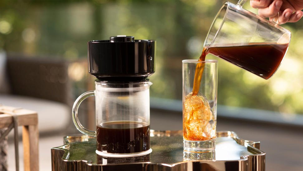 A VacOne Air Brewer coffee maker is in the center of the image. Right next to it, a person is pouring coffee into a glass half-filled with ice cubes.