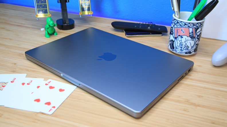 The Apple MacBook Pro 14 (2023) is sitting on a table, closed, next to a handful of playing cards, a cup filled with pens, and a green, toy dinosaur.