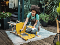 Person painting vintage chair with yellow paint on a patio surrounded with plants