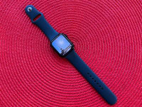 The Apple Watch SE with black band sits on a circular red background on a table.