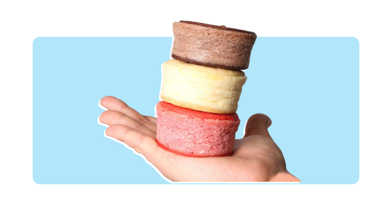 A hand holding three different colored cheesecake sandwiches in front of a background.,