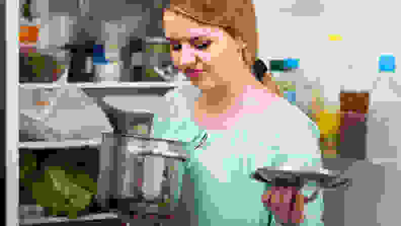 A woman grimaces at what's in a food storage container in front of an open fridge.