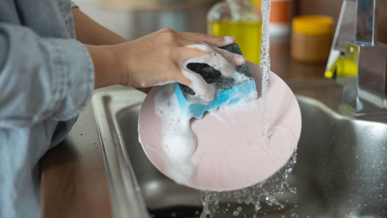 A person washing a plate in the sink with a soapy sponge and bare hands.