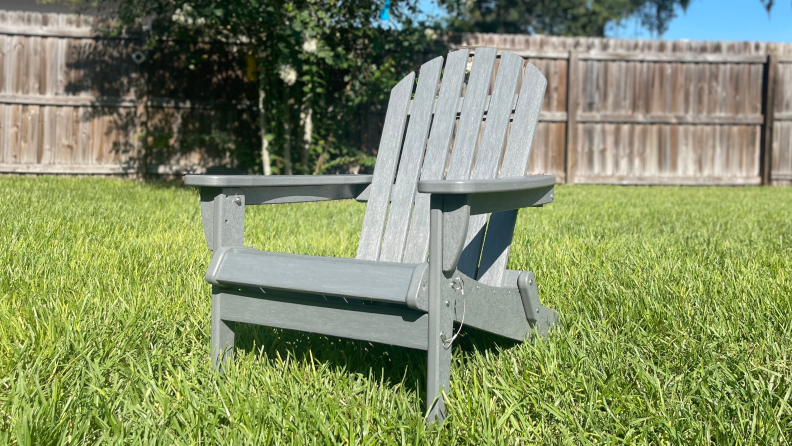 A gray Keter Willoughby Folding Adirondack Chair sitting in the grass with a fence and tree in the background