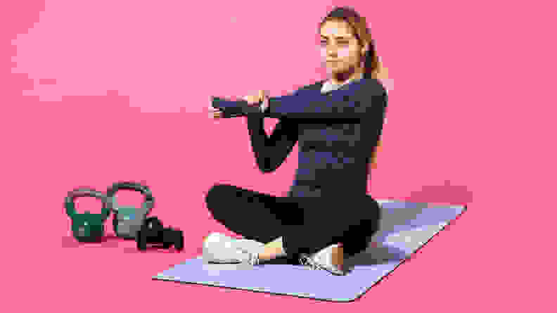 Person sitting cross-legged on Lululemon Workout Mat while stretching left arm across their body next to kettlebells.