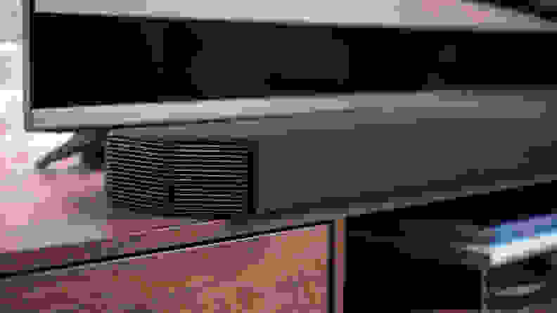 A close-up of the black Q950a sits on a woodgrain cabinet in front of a large TV, with brick background.