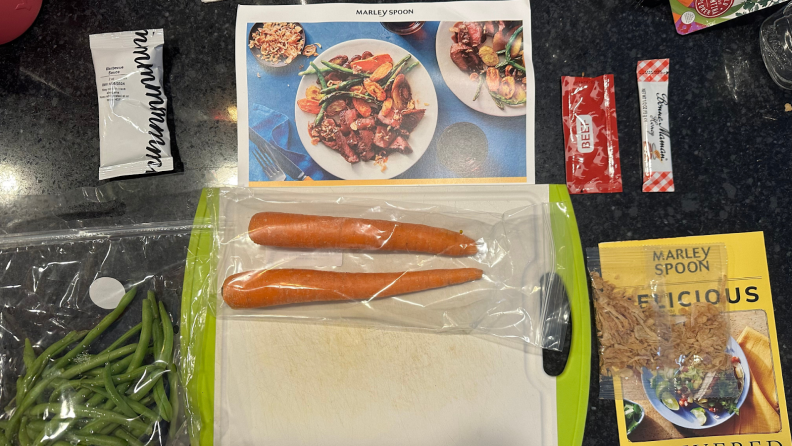 A bag of string beans next to two carrots on a cutting board below a Marley Spoon recipe card.