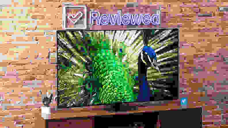 Samsung QN90D Mini-LED TV with vibrant peacock on screen atop a wooden TV stand below Reviewed neon sign in front of brick wall indoors.