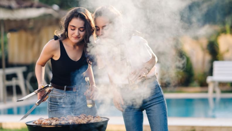 Two people grilling outdoors with a pool and backyard in the background.