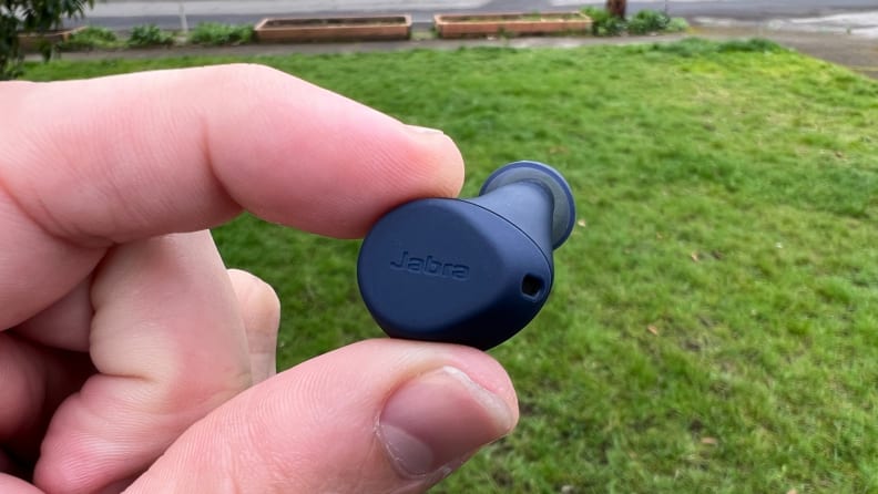 The navy blue Elite 7 Active earbud is held between thumb and forefinger in front of a green lawn with garden beds in the background.