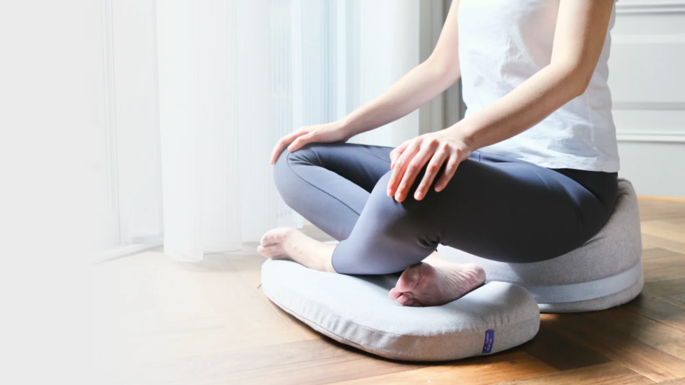 A person in yoga pants sits cross-legged on a chair cushion, which for the photo is positioned on a hardwood floor.
