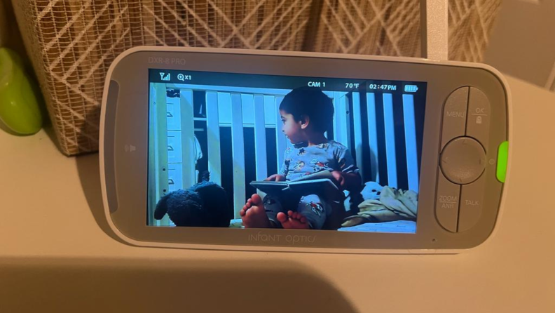 Small child looking off to the side with a book in their lap inside of crib on the screen of the Infant Optics DXR-8 Pro baby monitor.